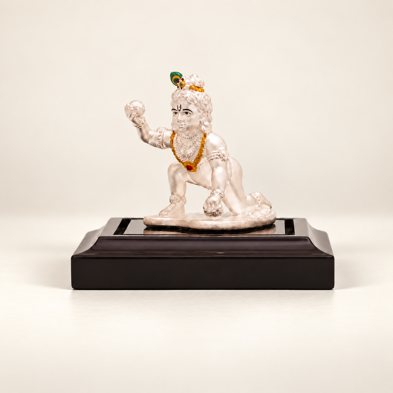 Silver crawling Laddu Gopal idol holding a sweet on a dark display stand against an off-white background.