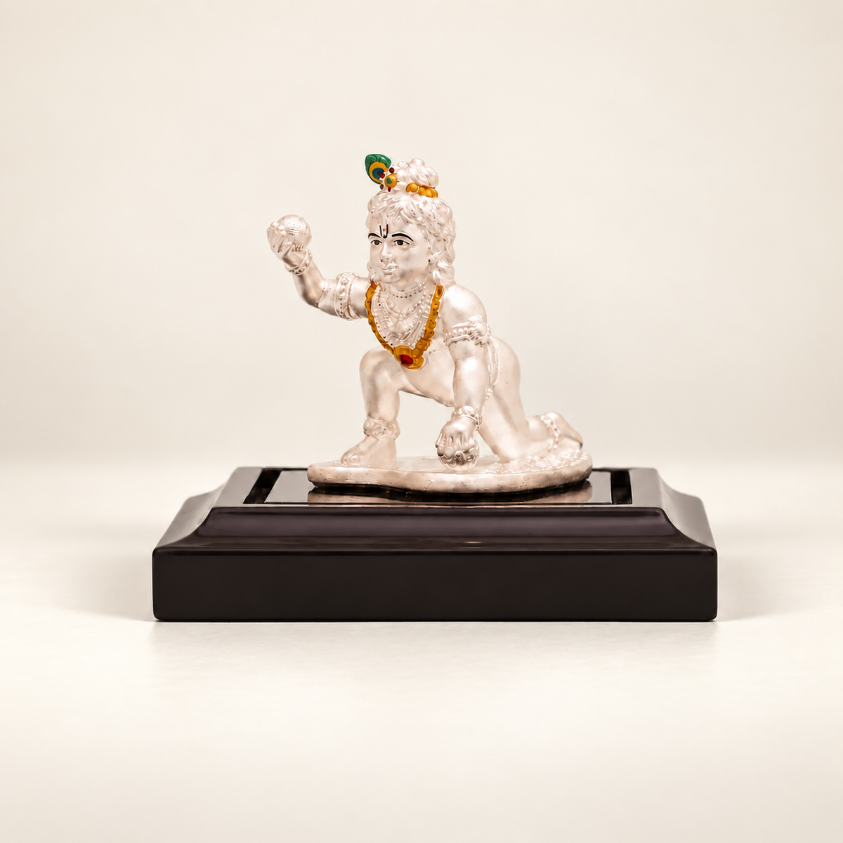 Silver crawling Laddu Gopal idol holding a sweet on a dark display stand against an off-white background.
