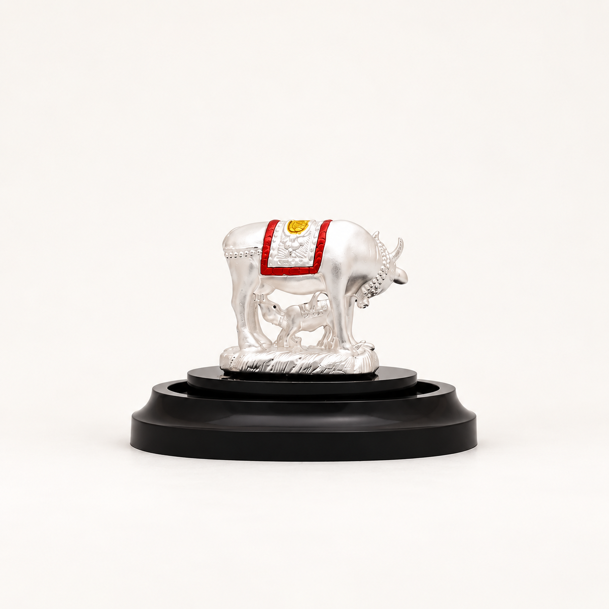 Side view of a silver cow and calf idol with red and gold detailing on a black pedestal against an off-white background.