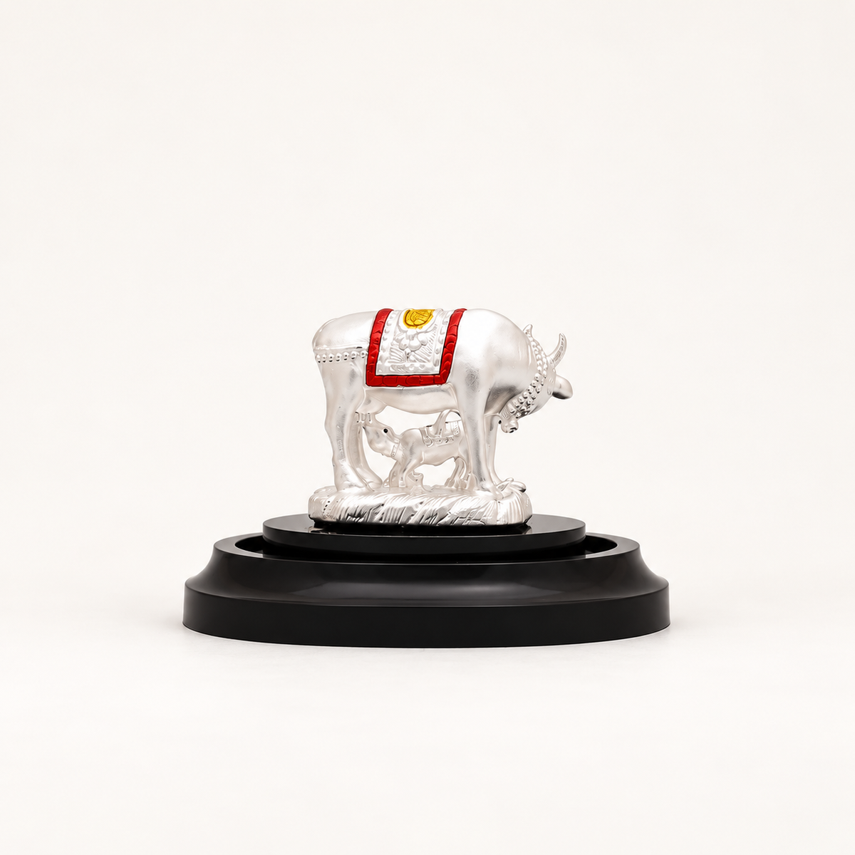 Side view of a silver cow and calf idol with red and gold detailing on a black pedestal against an off-white background.