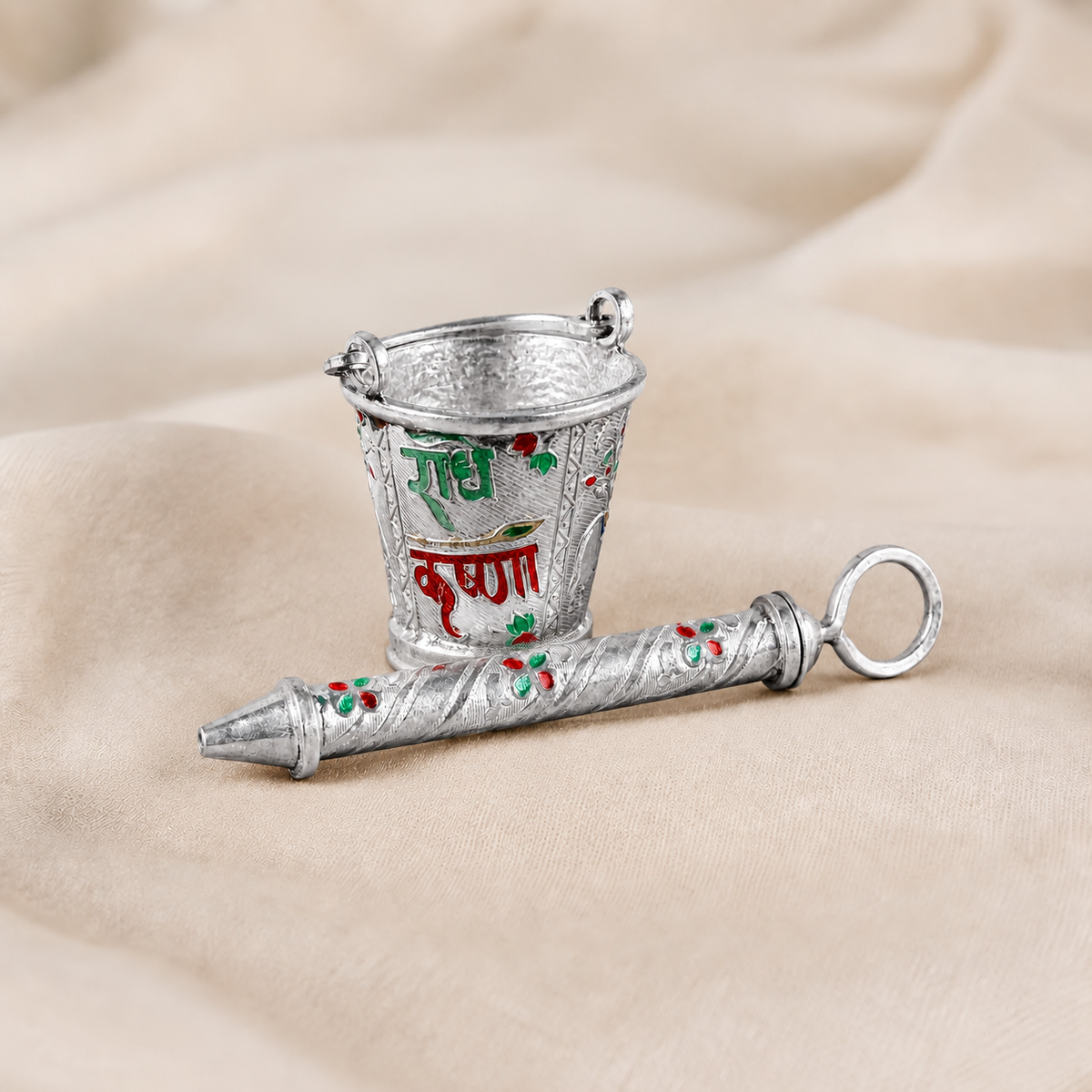 Engraved 925 silver bucket with green and red Radha Krishna lettering and matching floral pichkari on cream fabric.