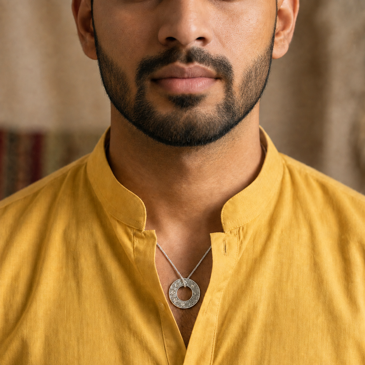 A close-up of a man wearing a silver-toned circular pendant necklace with a textured outer ring and a smooth center, styled with a mustard yellow shirt.
