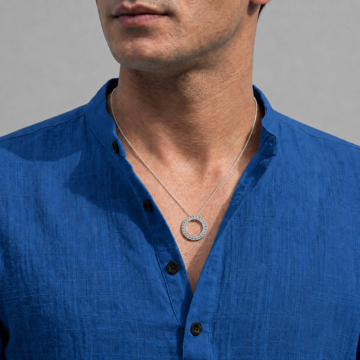 A close-up of a man wearing a silver circular pendant necklace with a halo of small clear stones, paired with a textured blue linen shirt against a neutral gray background.
