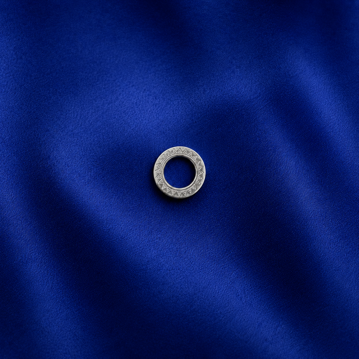 A silver-toned circular ring with a textured, patterned outer edge, photographed on a rich, dark blue satin fabric background.