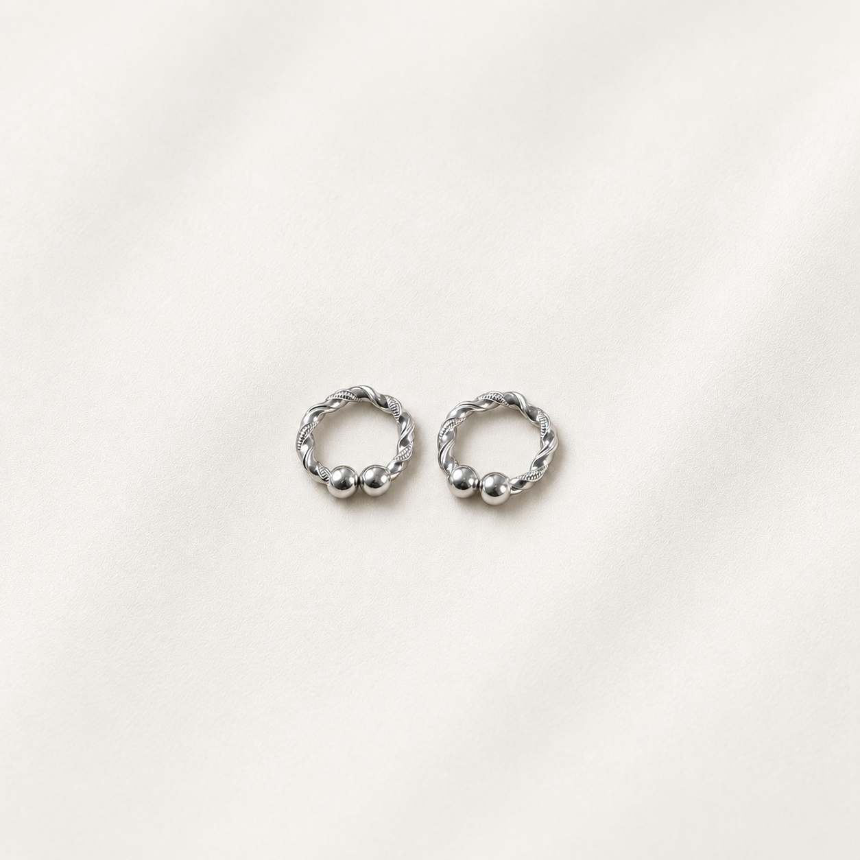 A pair of small silver hoop earrings with twisted rope detailing and small bead accents, displayed on a soft off-white fabric background.