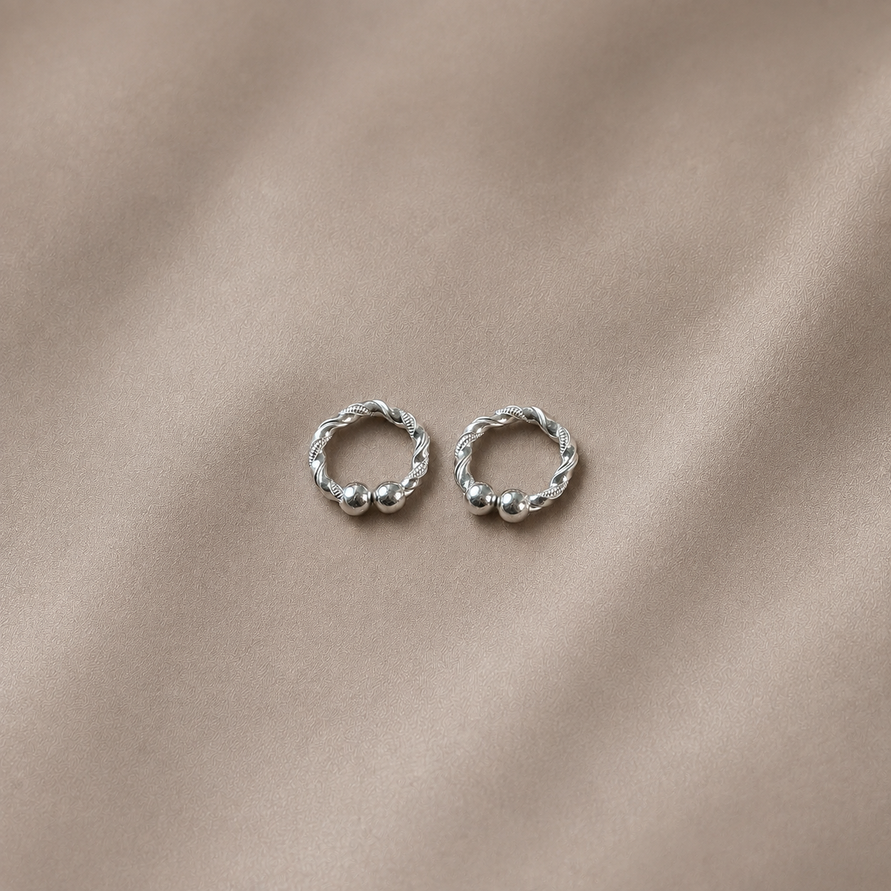 A pair of small silver hoop earrings with twisted rope detailing and small bead accents, displayed on a soft beige fabric background.