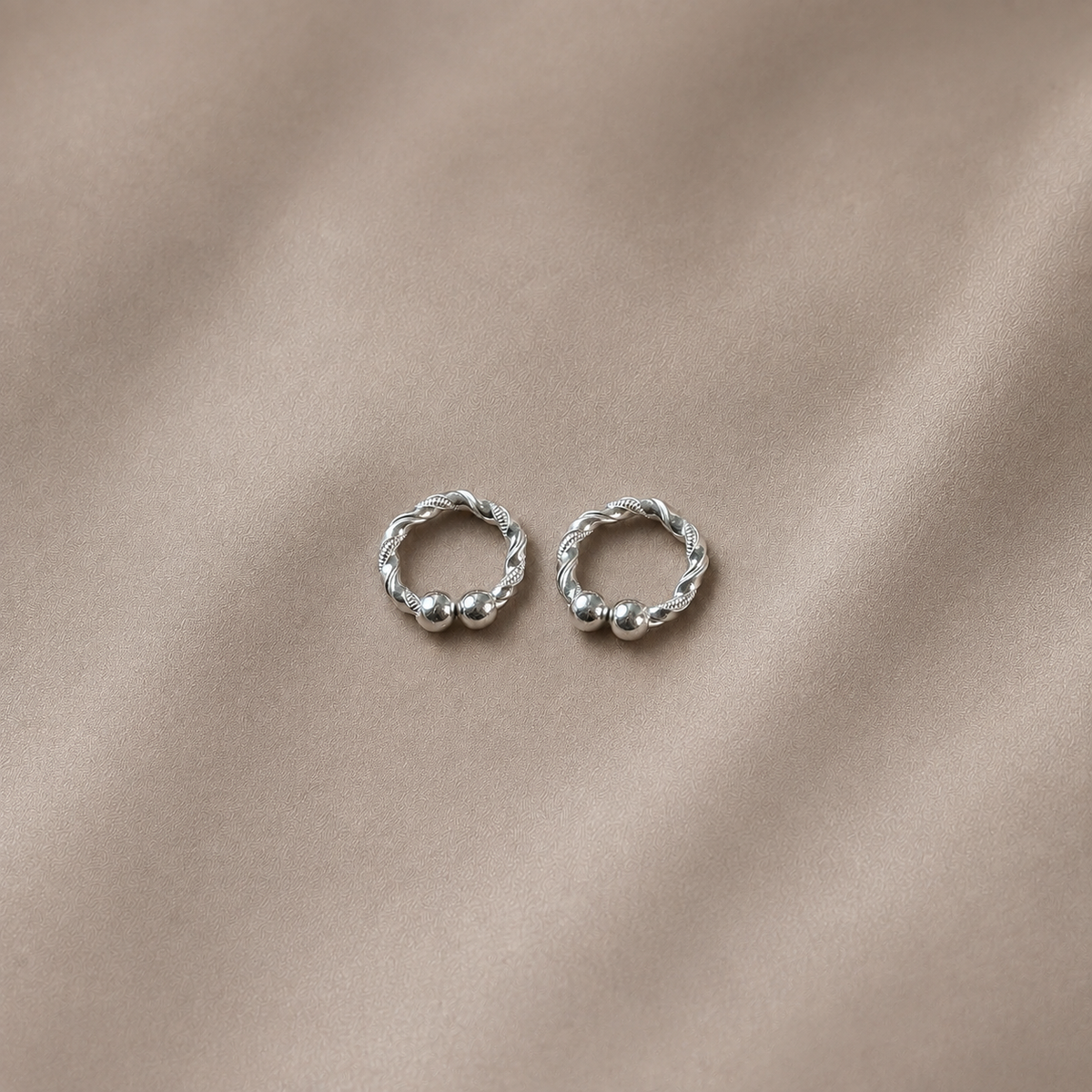 A pair of small silver hoop earrings with twisted rope detailing and small bead accents, displayed on a soft beige fabric background.