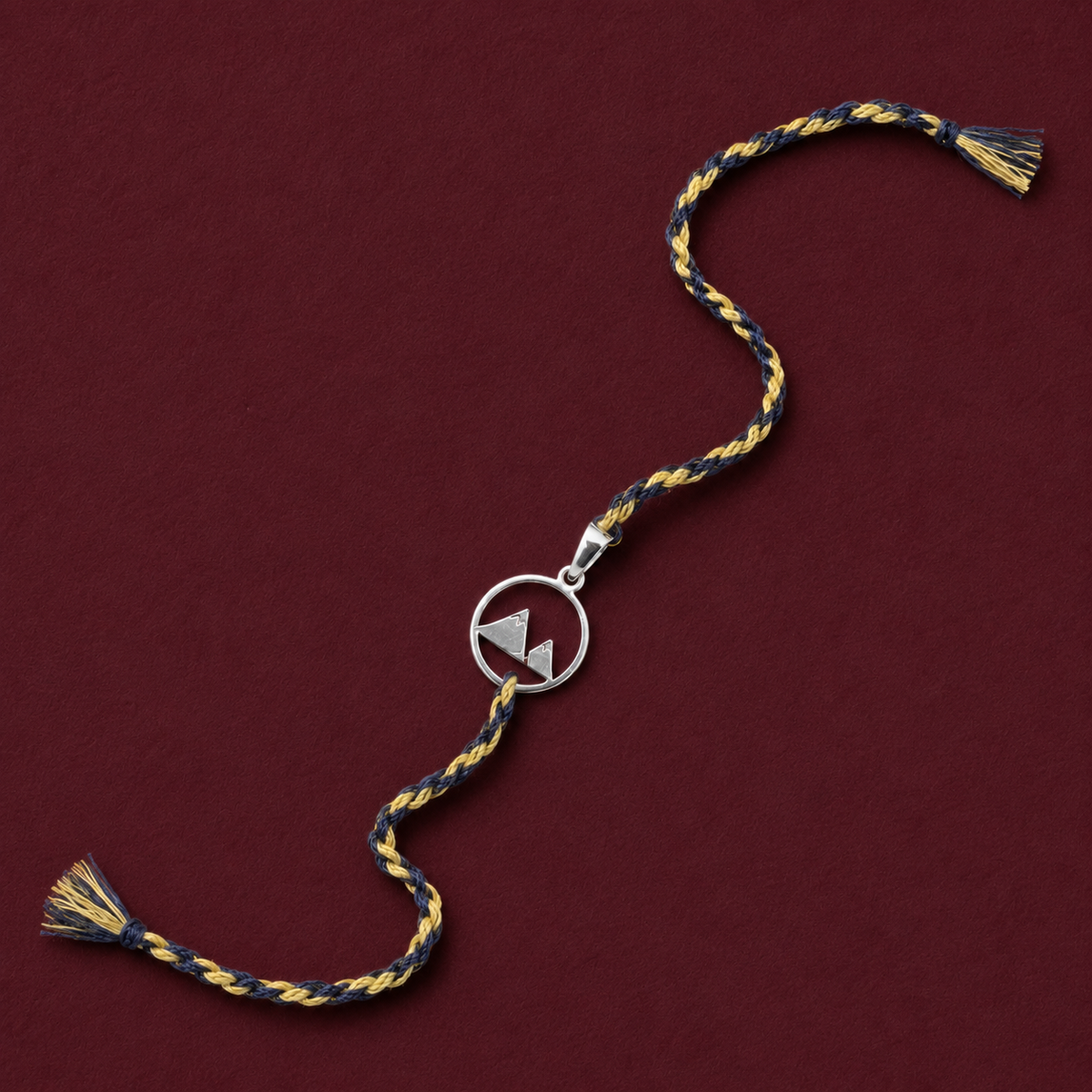 Pure silver mountain peak rakhi with grey and gold braided thread on dark red background