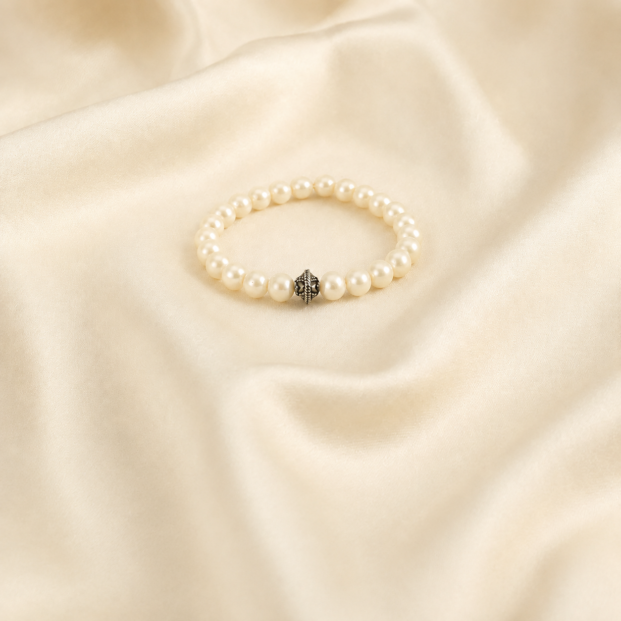 A beaded stretch bracelet with white faux pearls and a central silver-toned geometric charm, displayed on draped cream satin fabric.