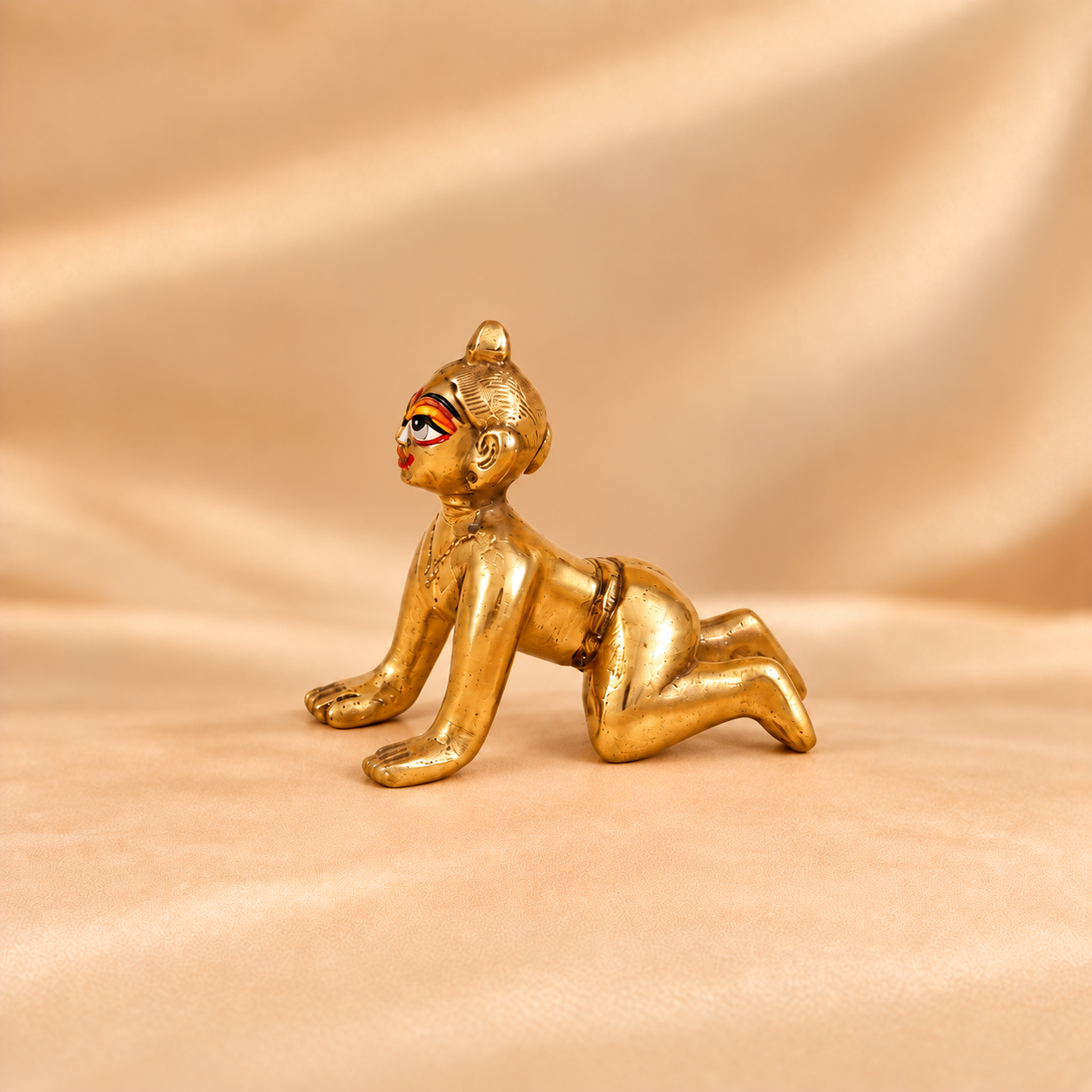 A small brass figurine of Radha Rani with stylized red and black facial makeup, posed on a beige satin-like fabric background.