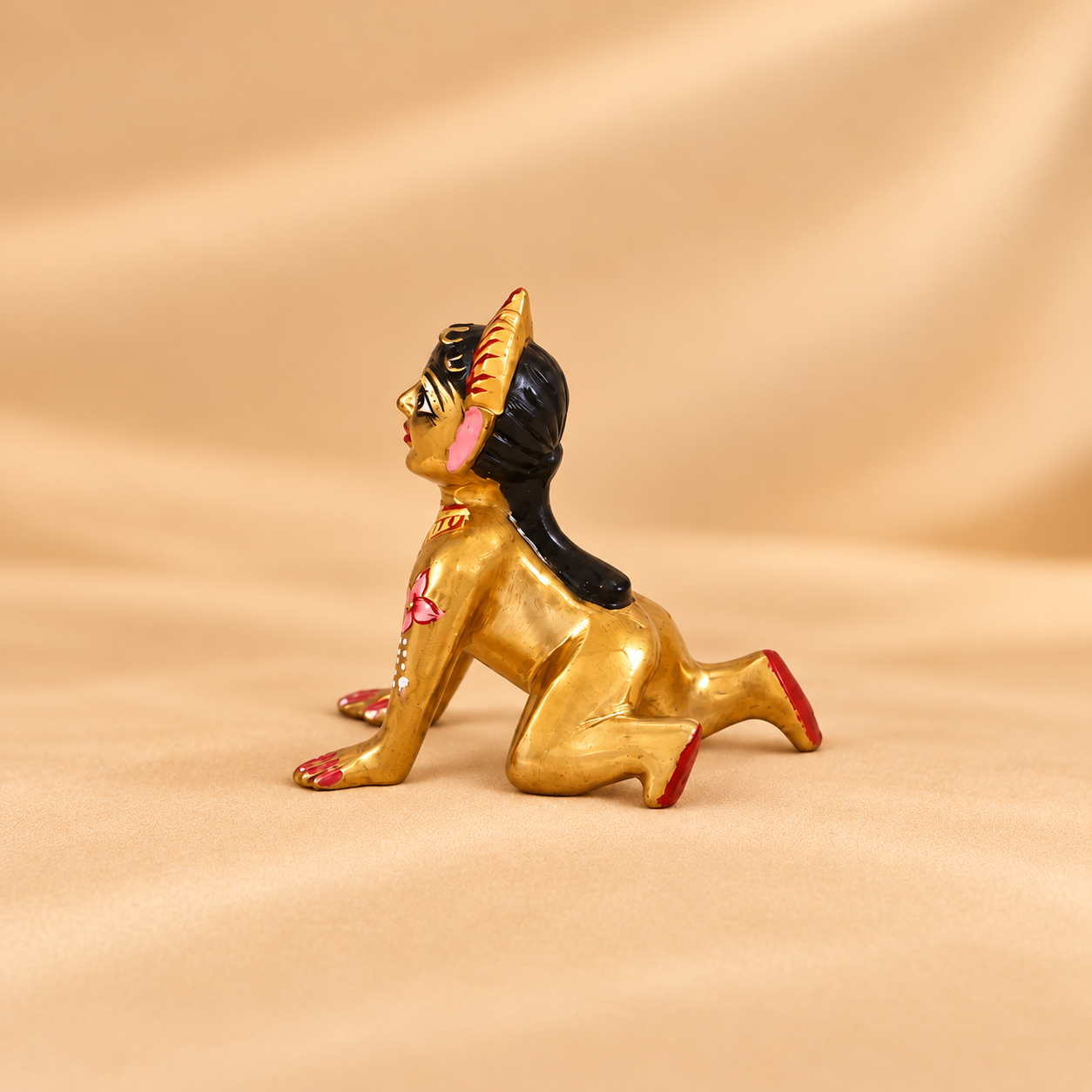 A small golden color figurine of a kneeling woman with black hair, pink details, and red accents, posed on a draped beige fabric background.
