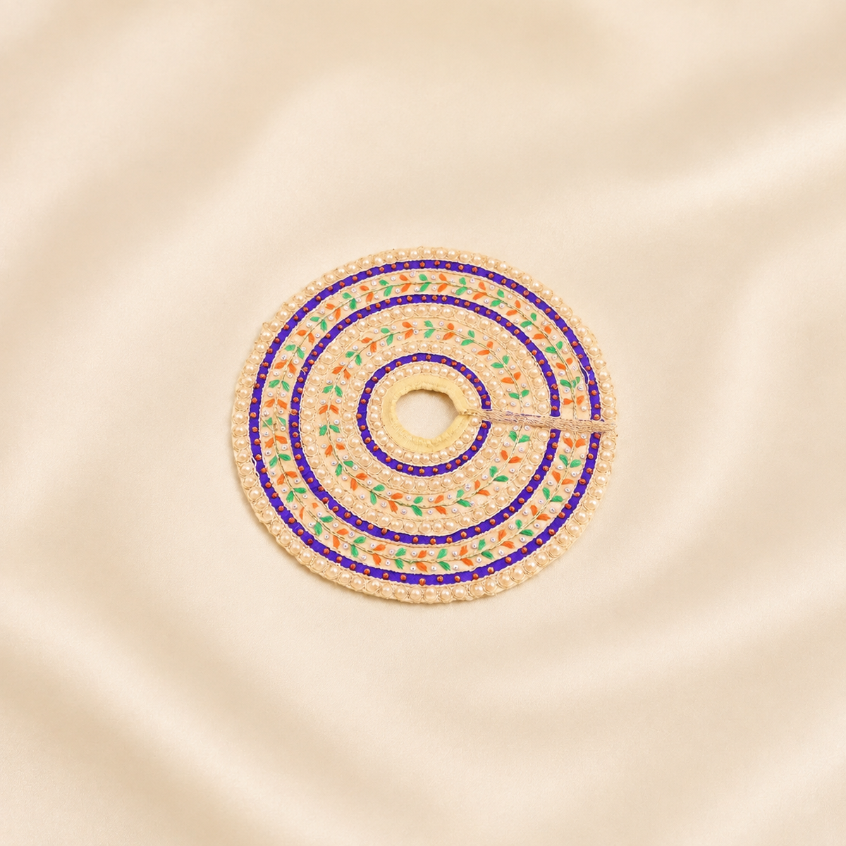 A circular embroidered poshak with concentric rings of white beads and colorful floral patterns in blue, green, and orange, displayed on a beige satin fabric background.