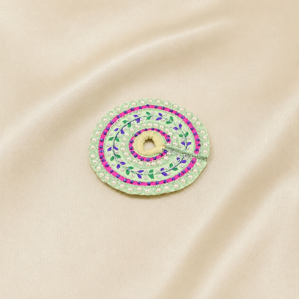 A circular embroidered poshak with a central hole, featuring green and purple floral patterns and a pink border, displayed on a beige satin fabric background.
