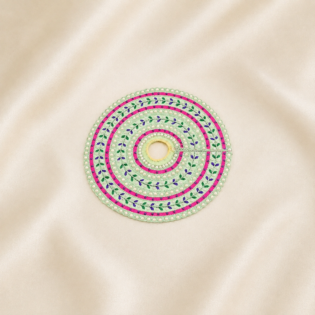A circular embroidered poshak with concentric rings of white pearls, bright pink thread, green and purple leaf motifs, and a central hole, displayed on a beige satin fabric background.