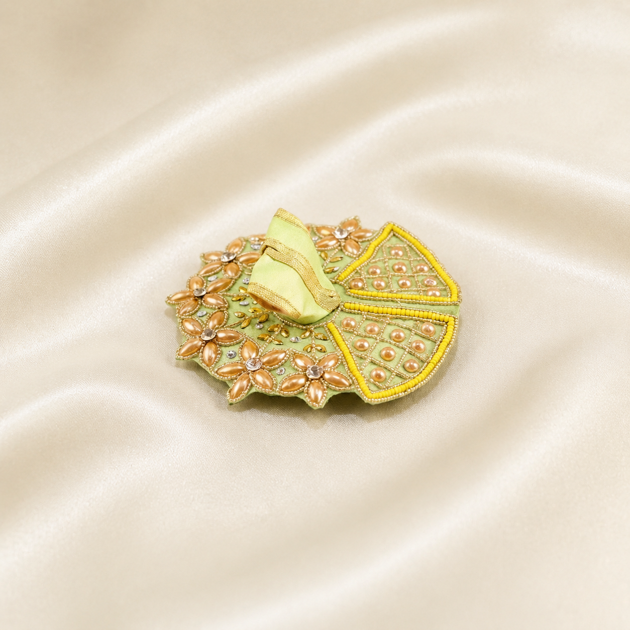 An ornate gold and green embroidered brooch shaped like a folded poshak, featuring gold-tone metalwork, green sequins, and peach-pink floral accents, displayed on a draped cream satin fabric background.