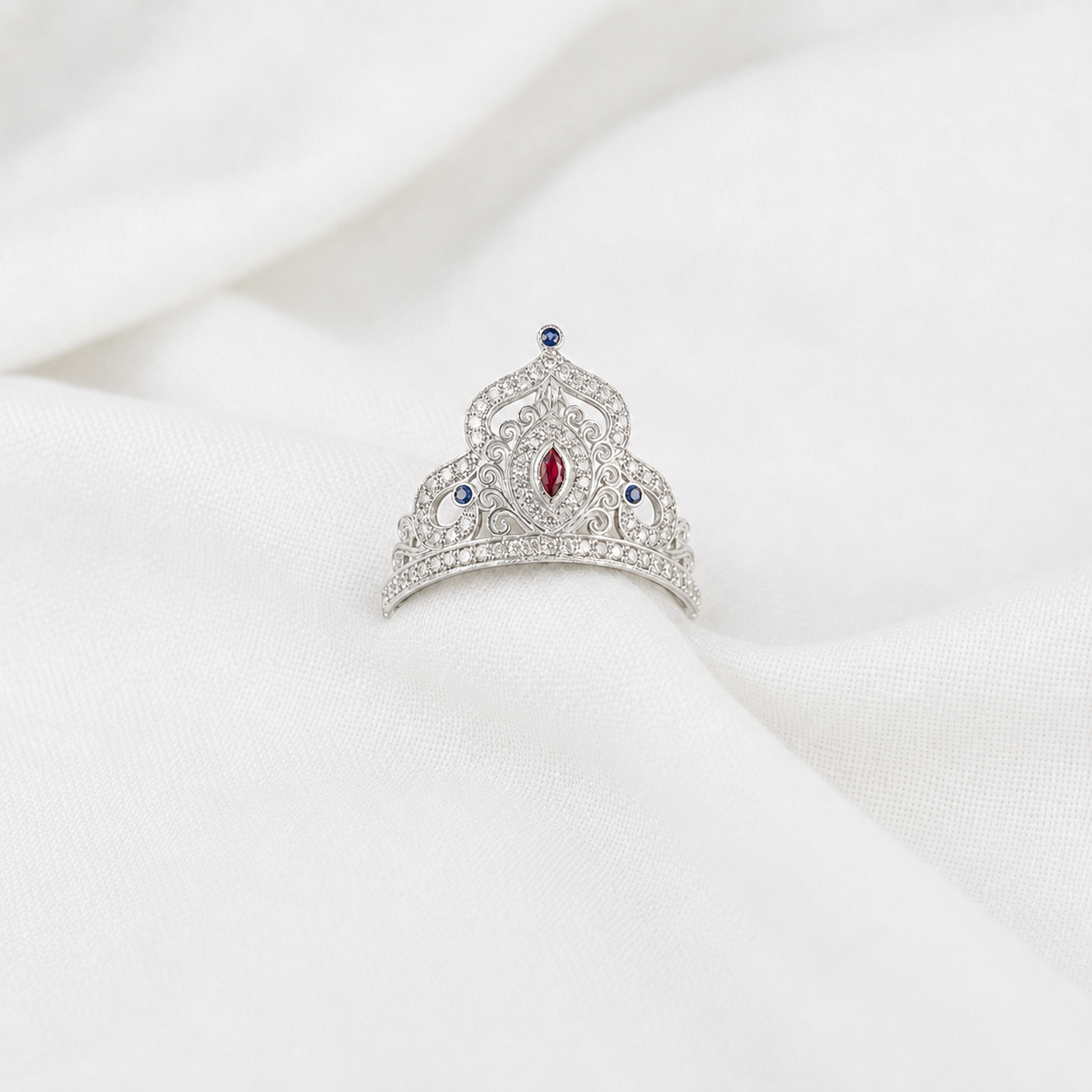 A silver mukut with a central red marquise-cut gemstone and small blue round gemstones, displayed on a draped white fabric background.