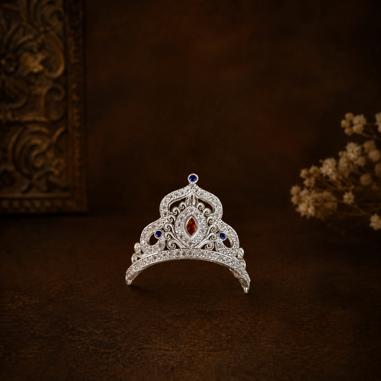 A silver mukut adorned with clear crystals, a central red gemstone, and blue gemstones, displayed on a dark brown textured surface with decorative background elements.