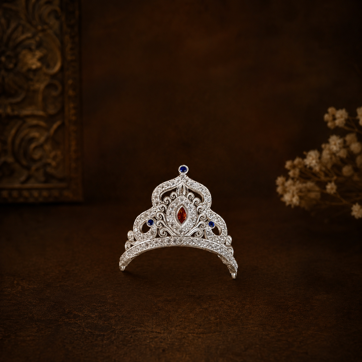 A silver mukut adorned with clear crystals, a central red gemstone, and blue gemstones, displayed on a dark brown textured surface with decorative background elements.