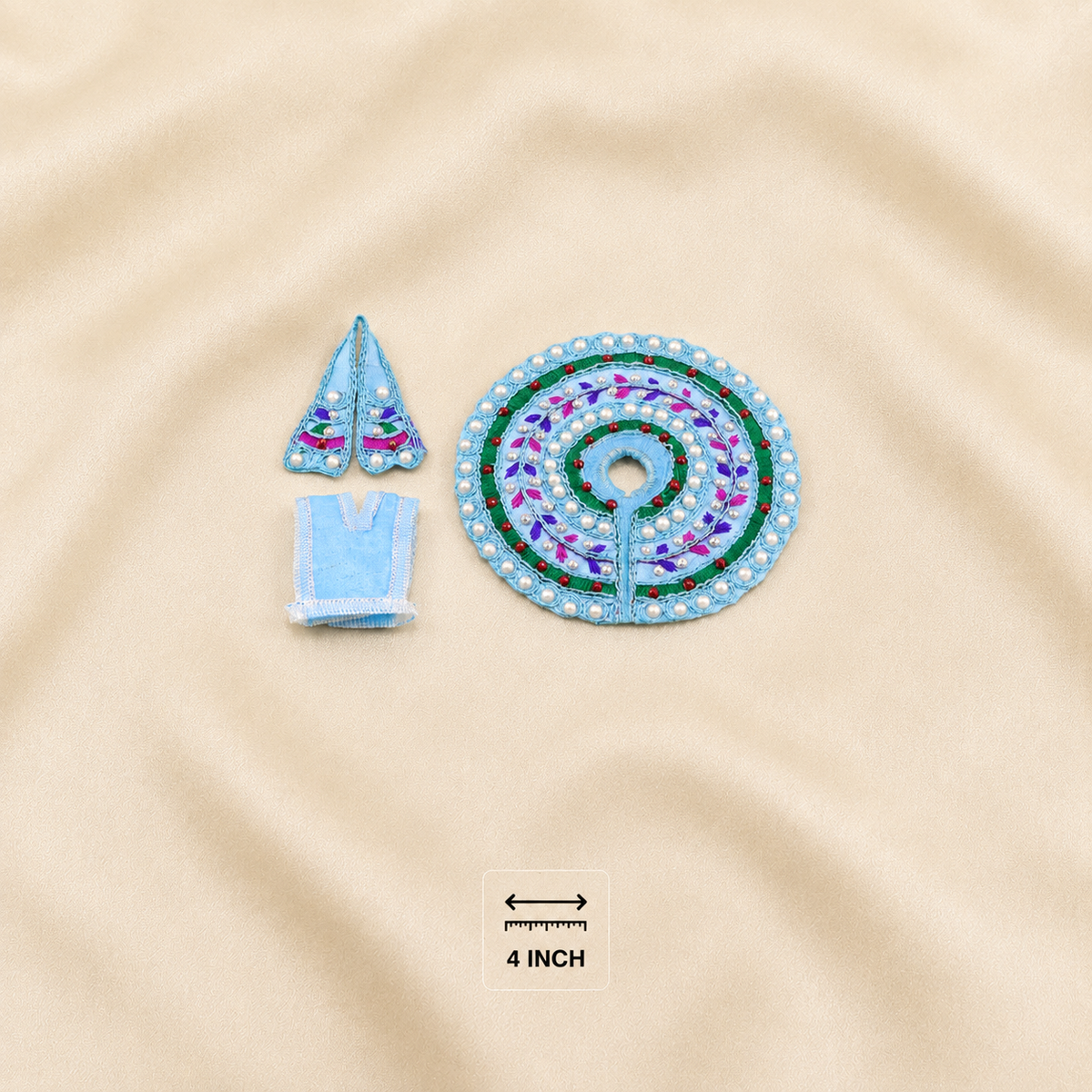 A small embroidered poshak set laid flat on a beige fabric background, including a circular patch with a spiral floral pattern, a small triangular embroidered piece, and a small rectangular piece. A small size marker reading 4 INCH is near the bottom.