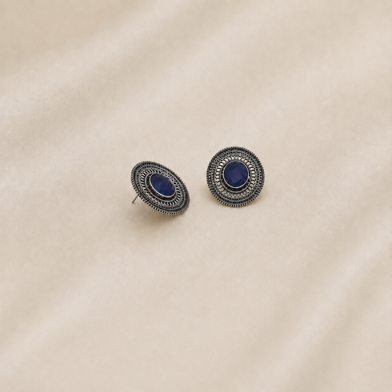 A pair of oval stud earrings with dark blue inlay and textured silver filigree borders, displayed on a soft beige fabric background.
