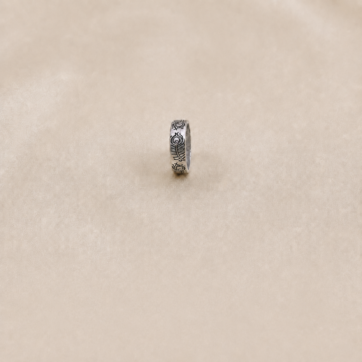 A silver ring with engraved floral or leaf-like patterns, standing upright on a textured beige surface.