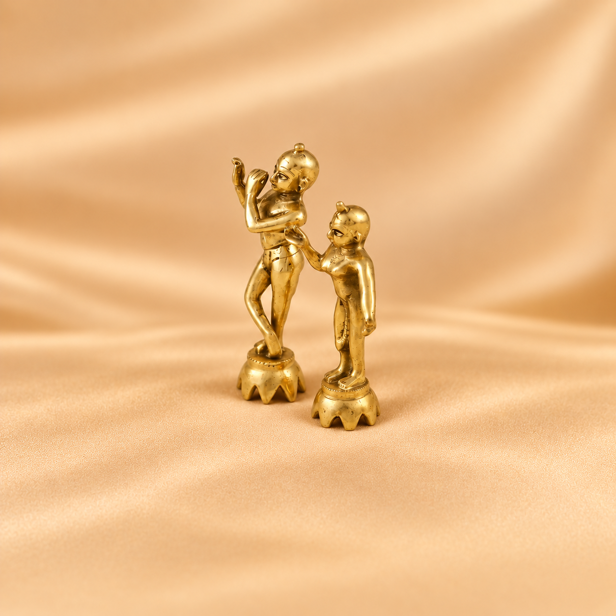 A small gold-toned figurine of radha krishna on decorative bases, photographed against a draped satin-like beige background.
