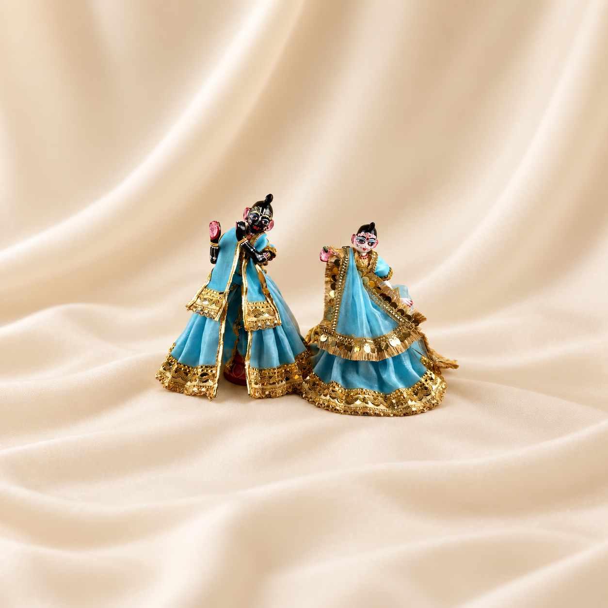 Radha Krishna idols wearing sky blue poshak with ornate gold-tone trim, placed together on cream fabric.