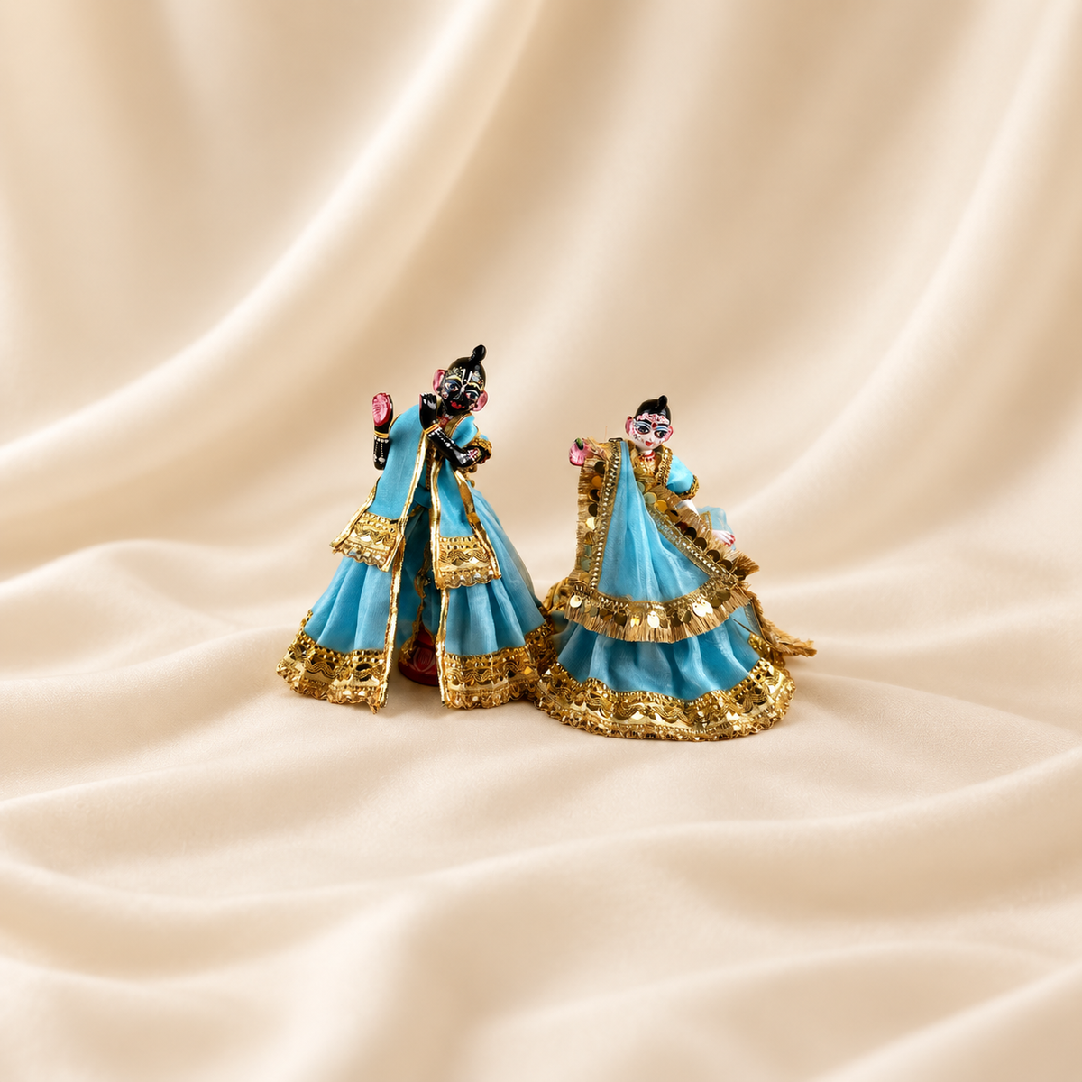 Radha Krishna idols wearing sky blue poshak with ornate gold-tone trim, placed together on cream fabric.
