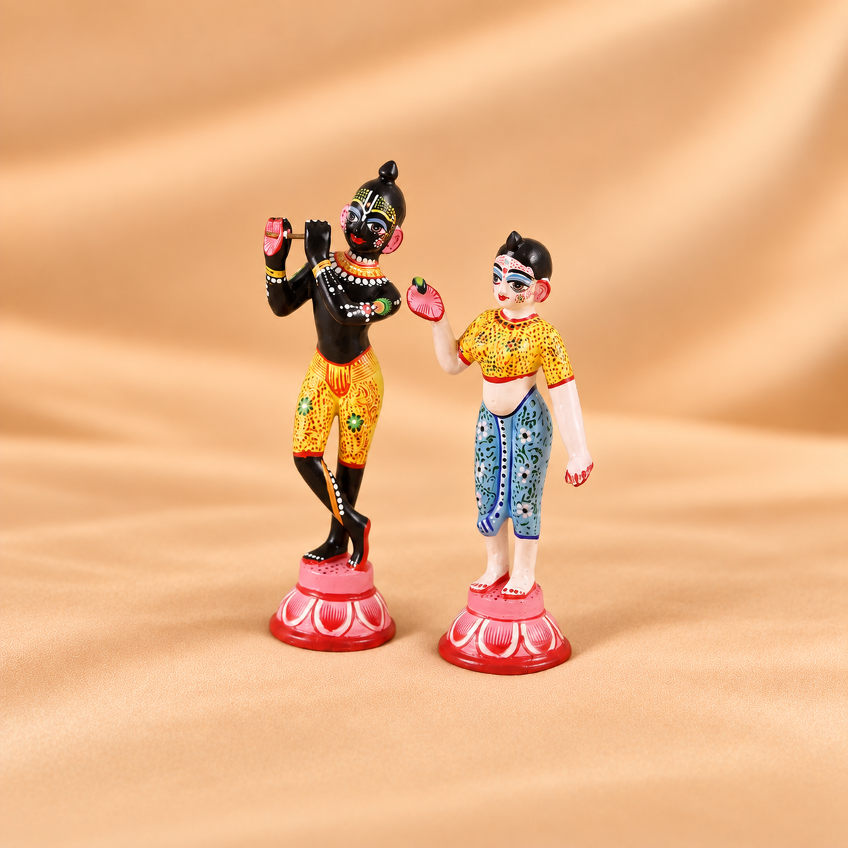 Handcrafted and hand-painted ashtadhatu Radha Krishna idols standing on pink lotus pedestals against a beige fabric background.