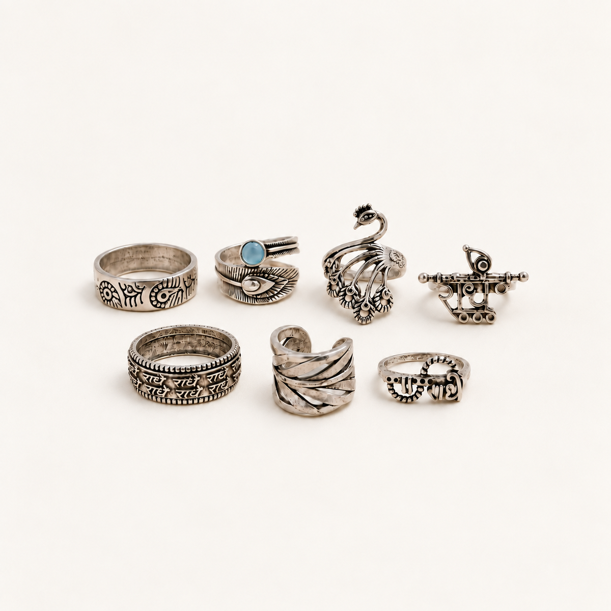 Seven silver-toned alloy rings with Radhe, peacock, feather and flute designs on an off-white background.