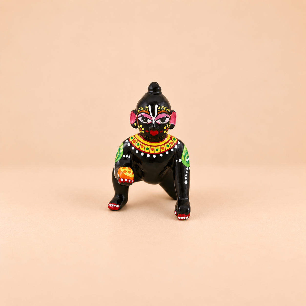 A small black laddu gopal with colorful painted facial features, a red nose, green and yellow shoulder details, and white dot patterns, posed against a solid beige background.