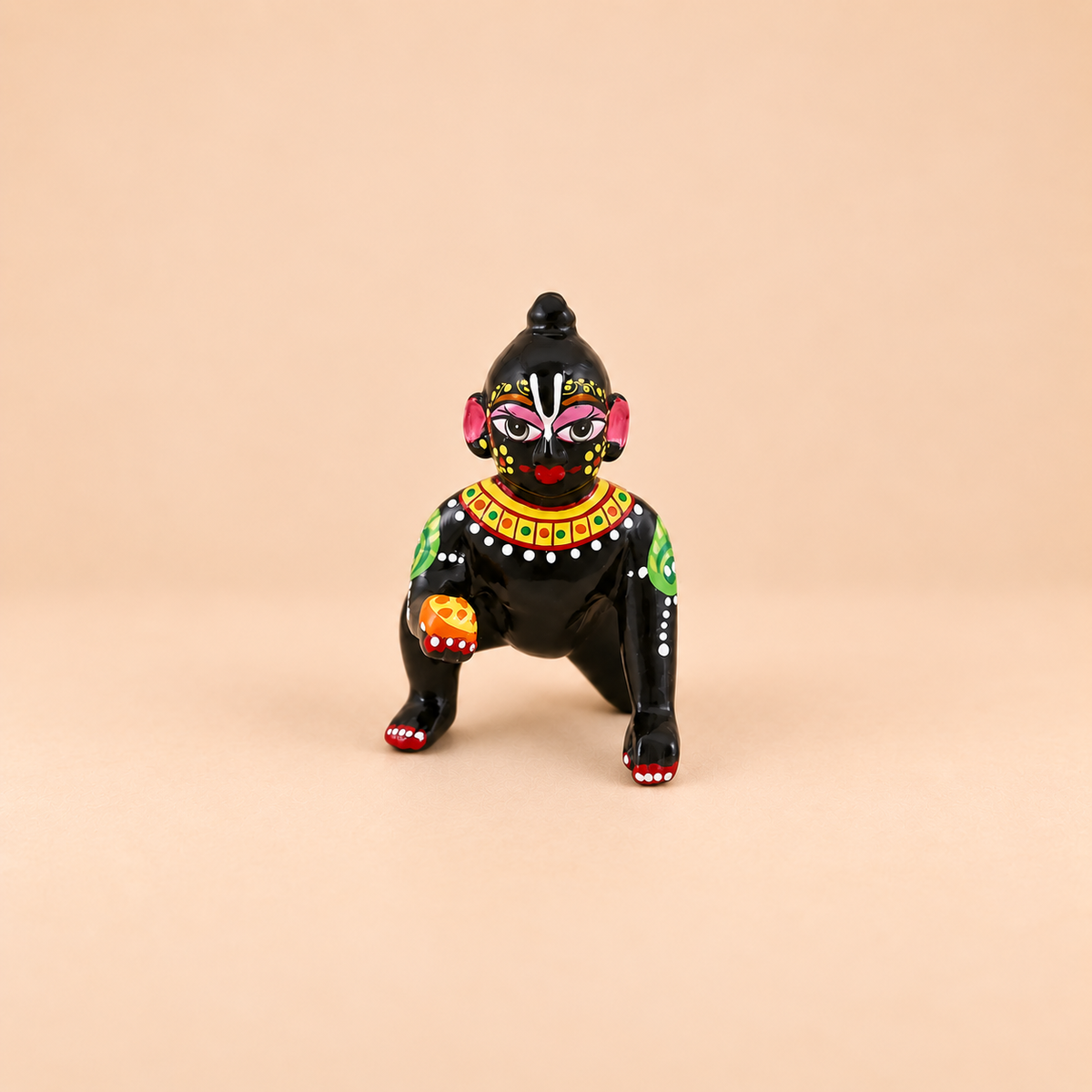 A small black laddu gopal with colorful painted facial features, a red nose, green and yellow shoulder details, and white dot patterns, posed against a solid beige background.