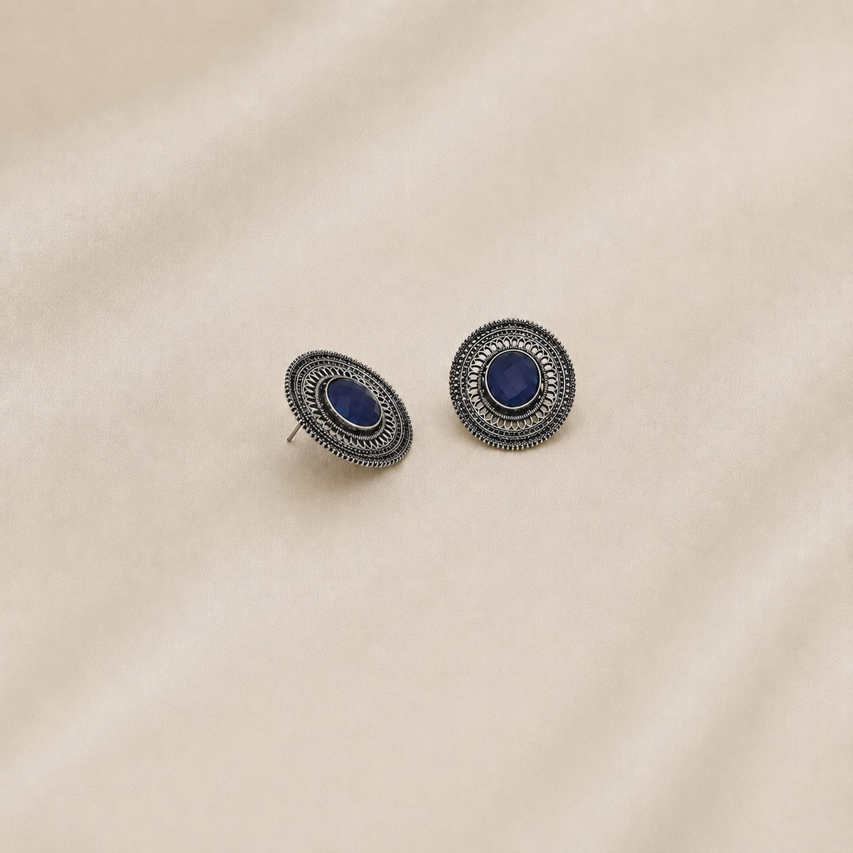 Pair of blue oxidized metal stud earrings with round filigree design displayed on beige fabric