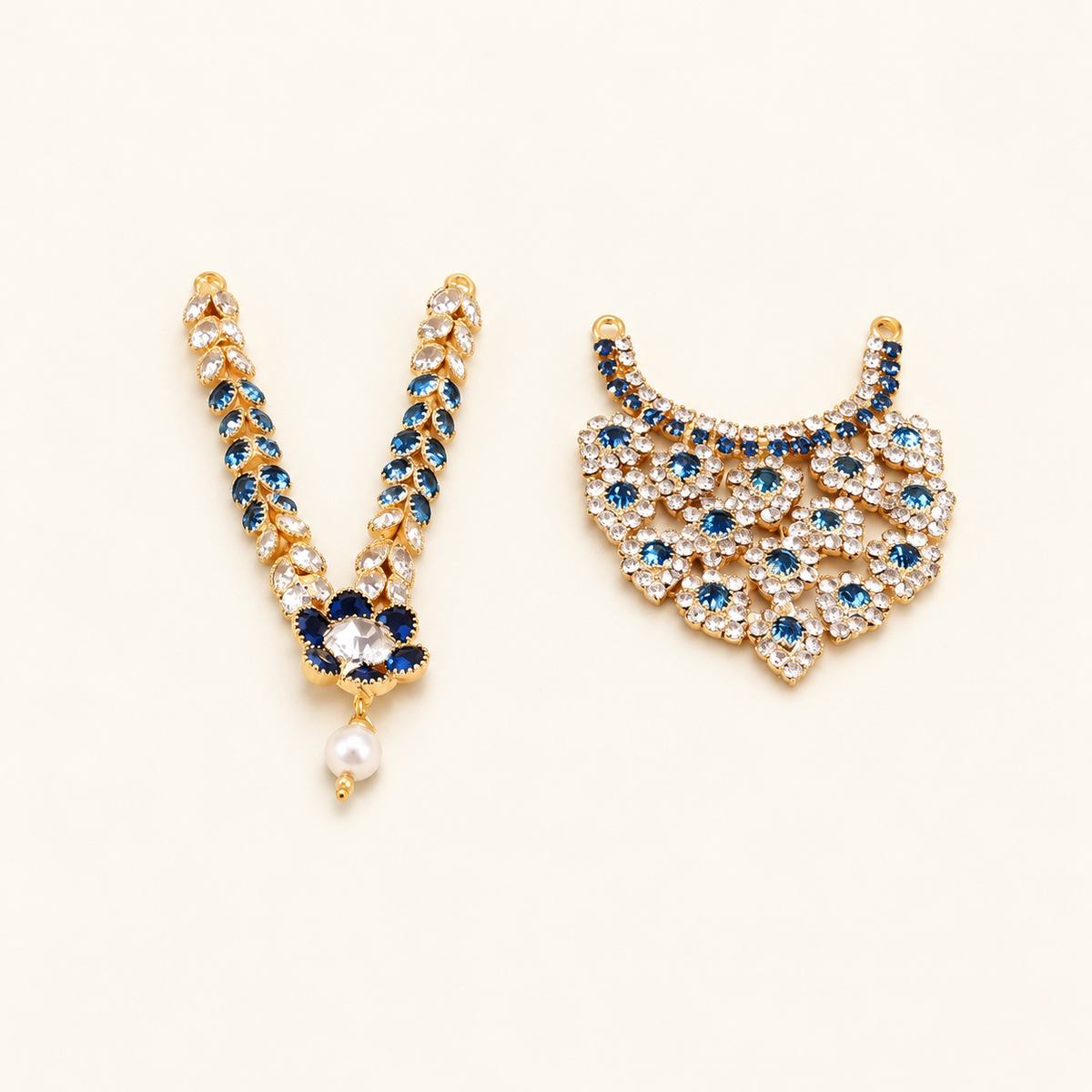 Two gold-toned imitation idol necklaces with blue and clear stones, one with a pearl drop, on an off-white background.