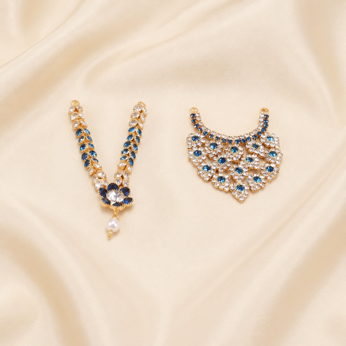 Two gold-toned imitation idol necklaces with blue and clear stones, one with a pearl drop, on cream satin.