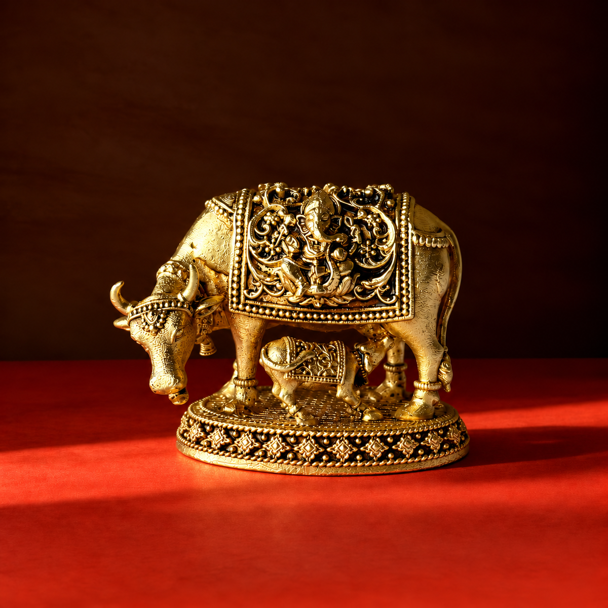 Brass Kamdhenu cow and calf statue with Ganesha relief and an ornate oval base on a red surface.