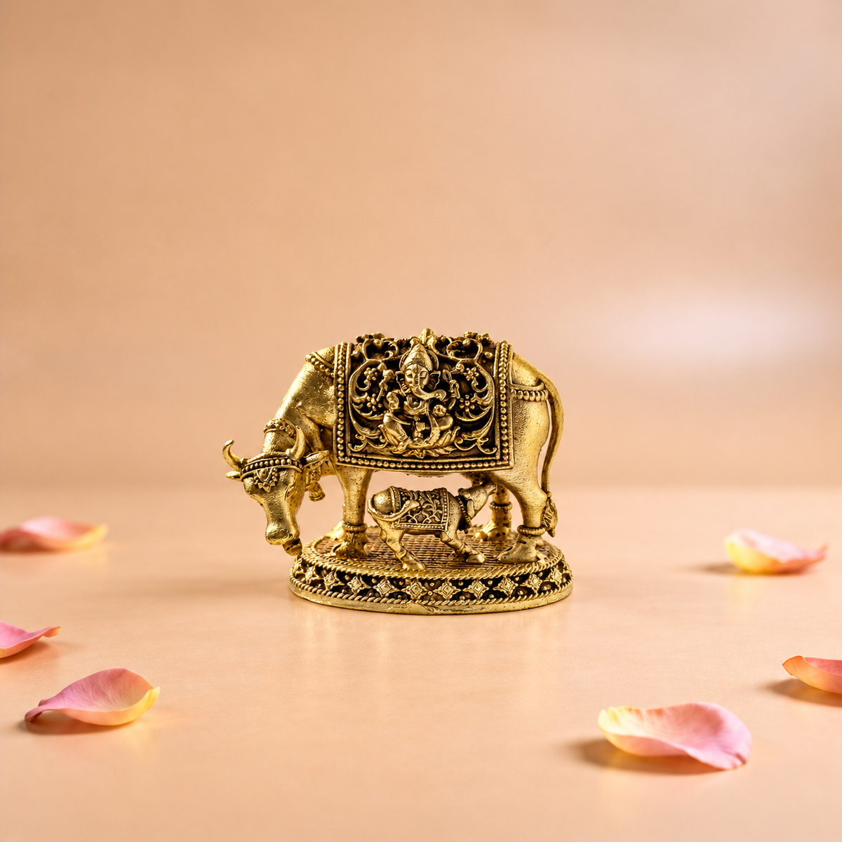 Golden brass Kamdhenu cow and calf statue with Ganesha relief on a peach background with scattered flower petals.