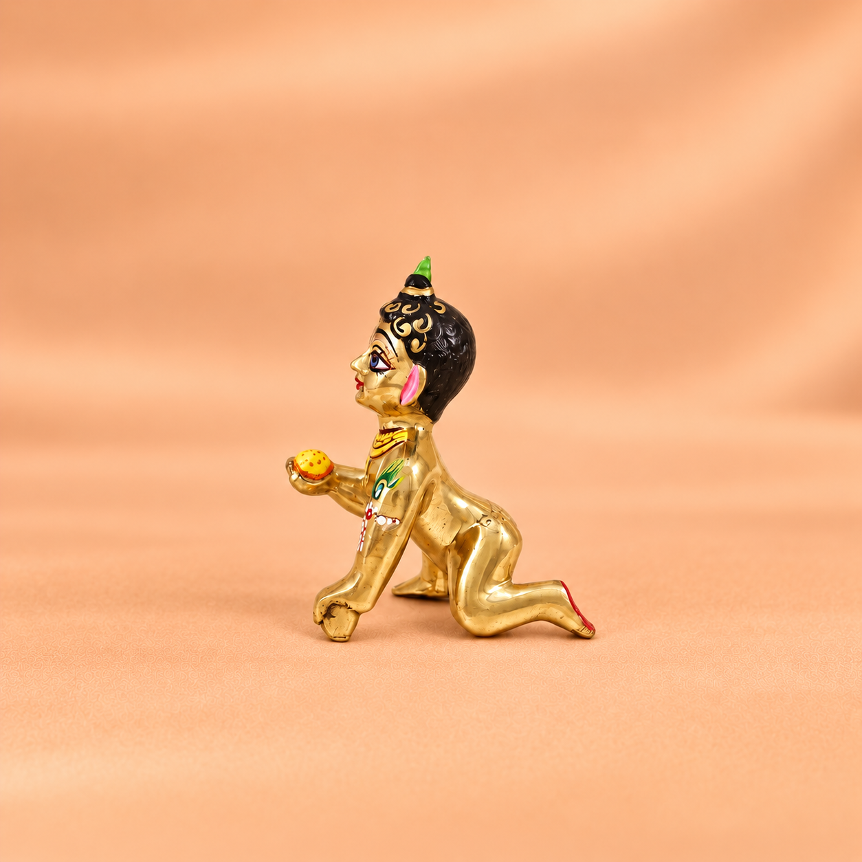 Side view of a brass Laddu Gopal idol holding a yellow laddu against a soft peach background.