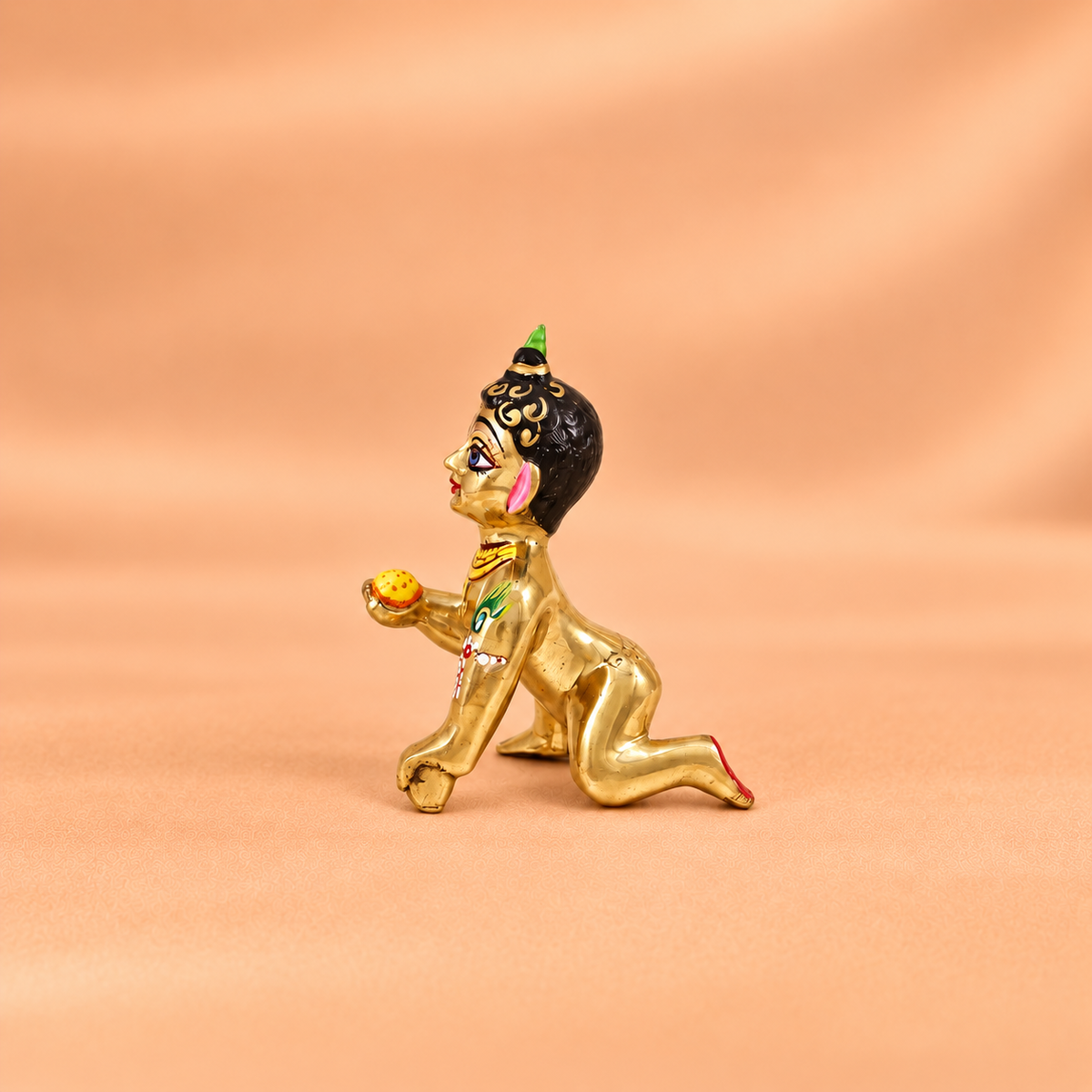 Side view of a brass Laddu Gopal idol holding a yellow laddu against a soft peach background.