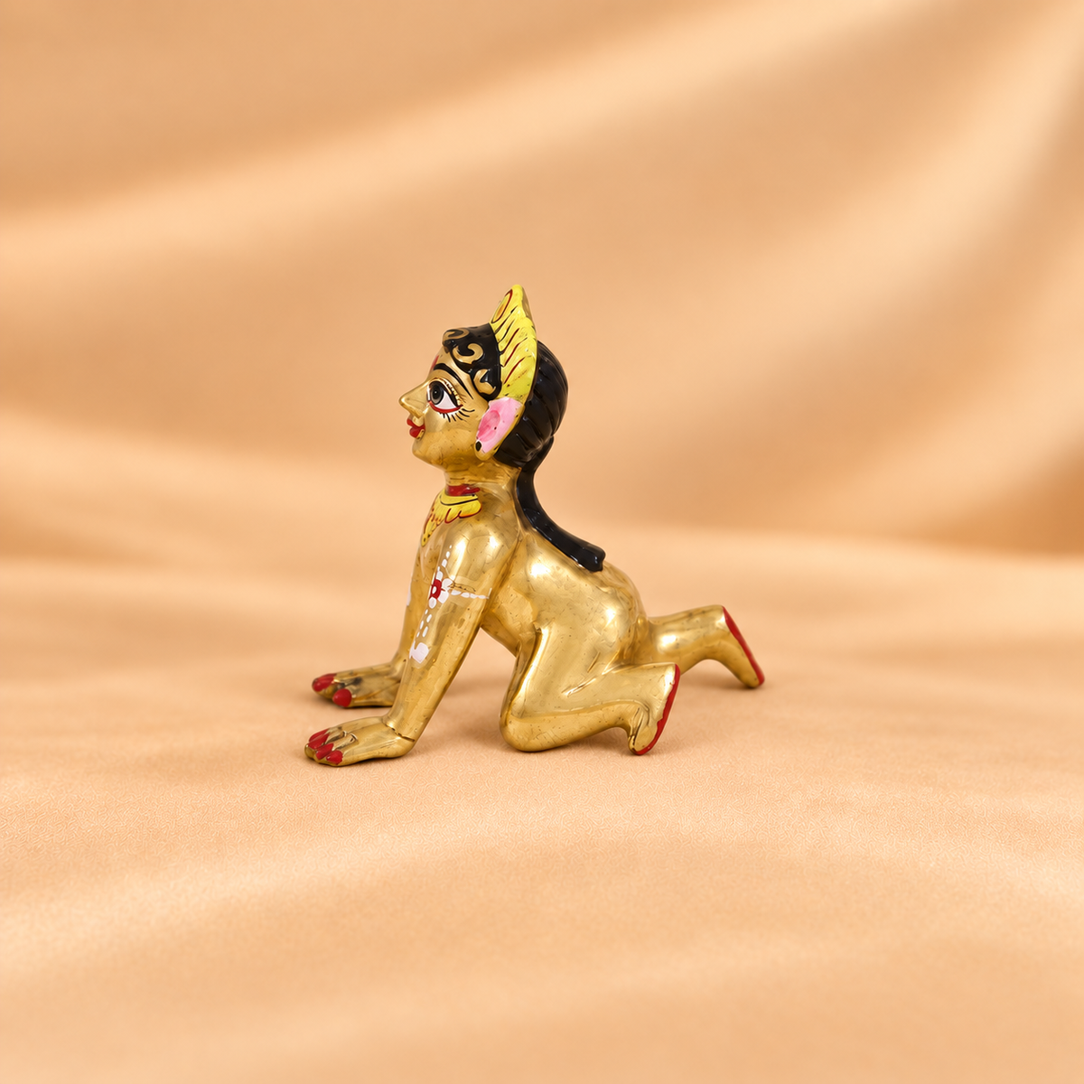 Side profile view of a brass Radha Rani crawling idol featuring detailed hand-painted body artwork on a soft beige silk background.