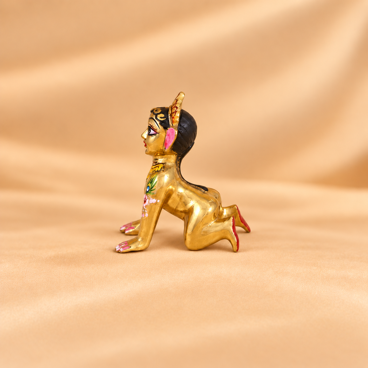 Side profile view of a brass Radha Rani crawling idol with detailed hand-painted painted artwork on beige fabric background.