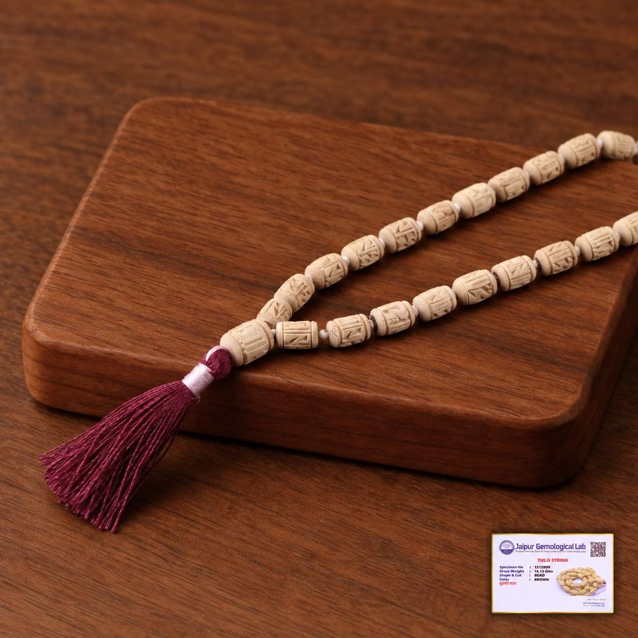 Carved Tulsi bead mala with a burgundy tassel on a wooden board, with a certificate displayed at the bottom right.
