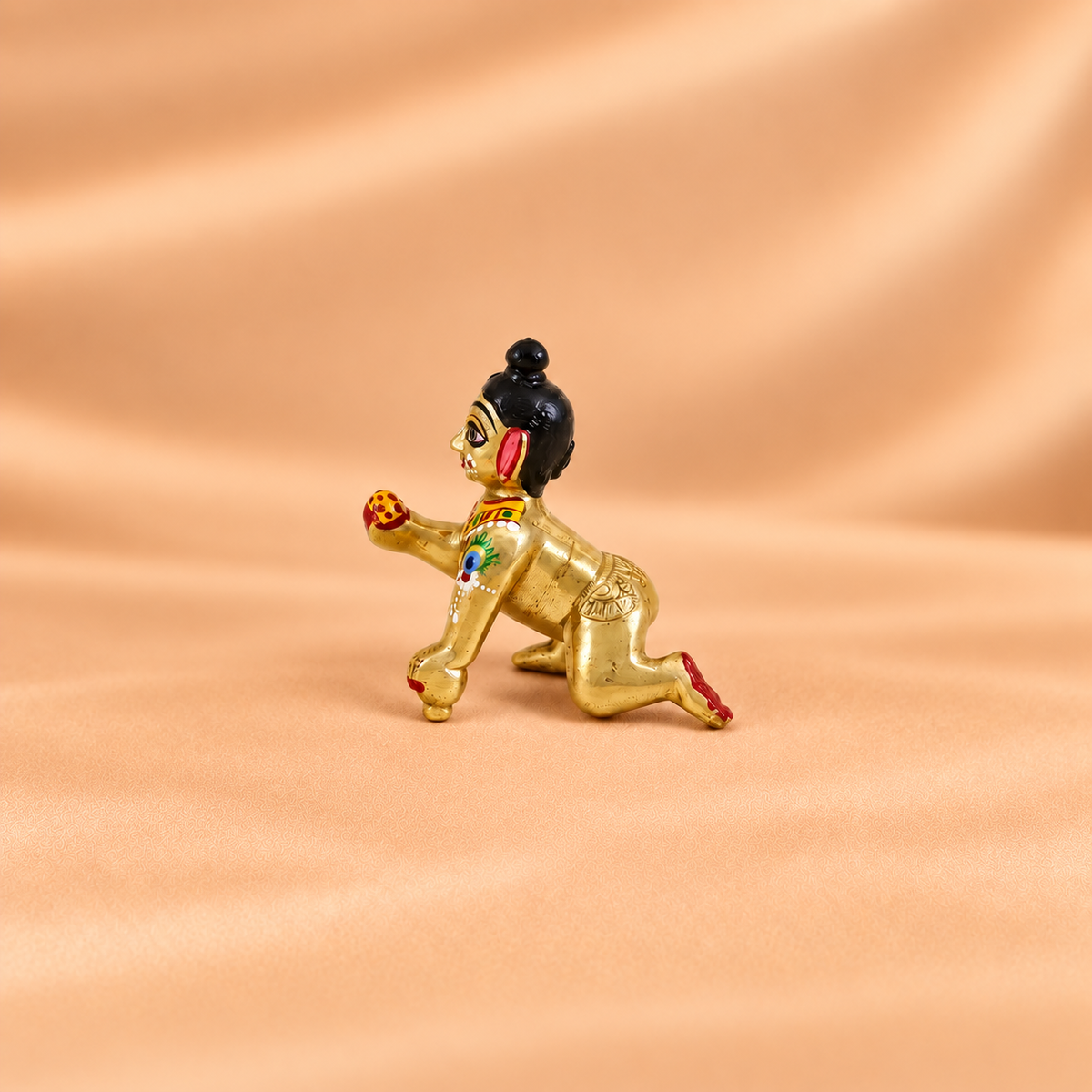 Side view of a golden crawling Laddu Gopal idol holding a laddu, with painted peacock feather detail on the arm