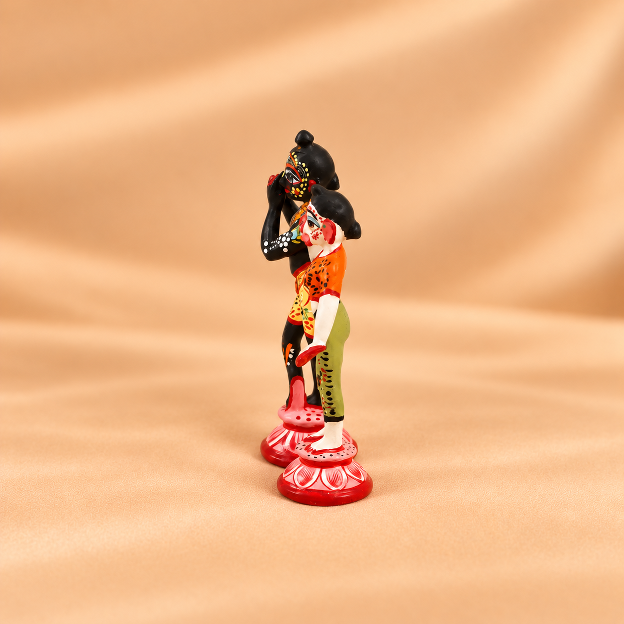 Side view of handcrafted and colorful hand-painted metal Radha Krishna idols standing on lotus pedestals against a beige fabric background.