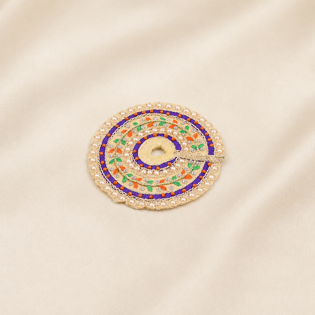 A circular embroidered poshak with concentric rings of gold, white pearls, blue, green, and orange thread, displayed on a beige satin fabric background.