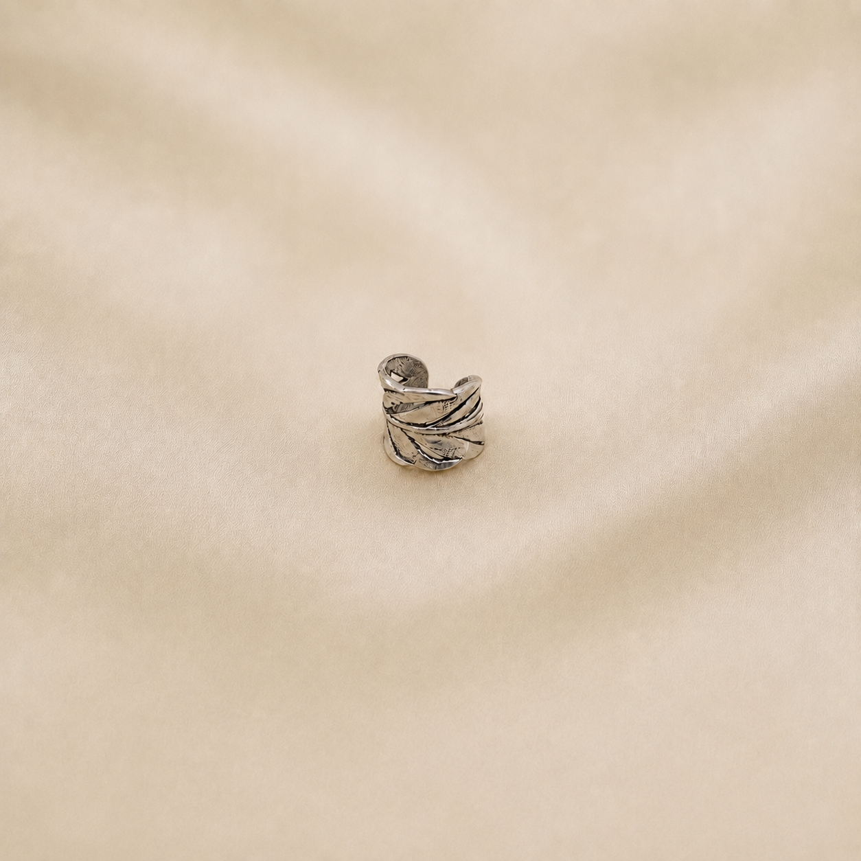 A small silver ring with a textured, leaf-like wrap design, displayed on a soft beige fabric background.