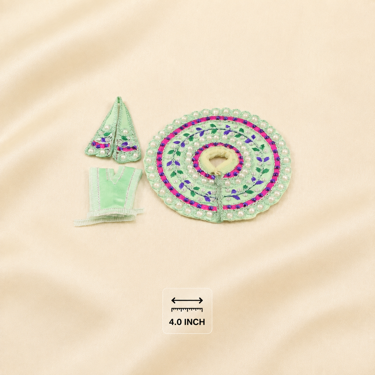 A miniature light green clothing set on a beige satin fabric background, featuring a circular skirt with pink and purple floral embroidery, a small pointed hat, and a small garment piece. A small scale marker indicating 4.