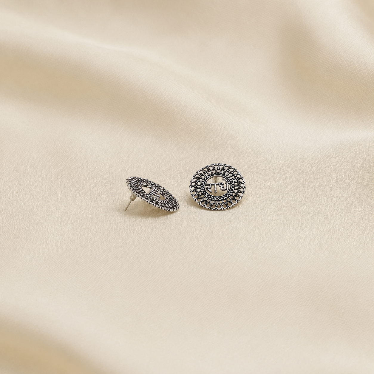 A pair of round silver-toned stud earrings with textured, beaded edges and a central fleur-de-lis motif, displayed on a draped beige fabric background.