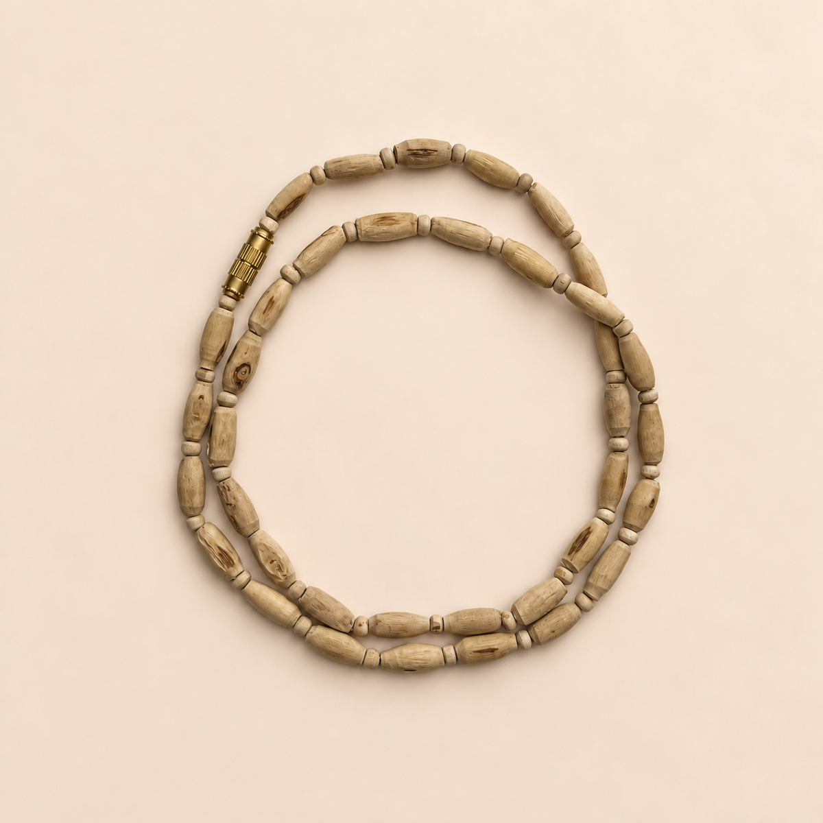 Natural Tulsi wood Kanthi Mala with elongated oval wooden beads, small round spacer beads, visible natural wood grain and a gold-tone screw clasp, arranged in a double circular loop on a pastel cream background.