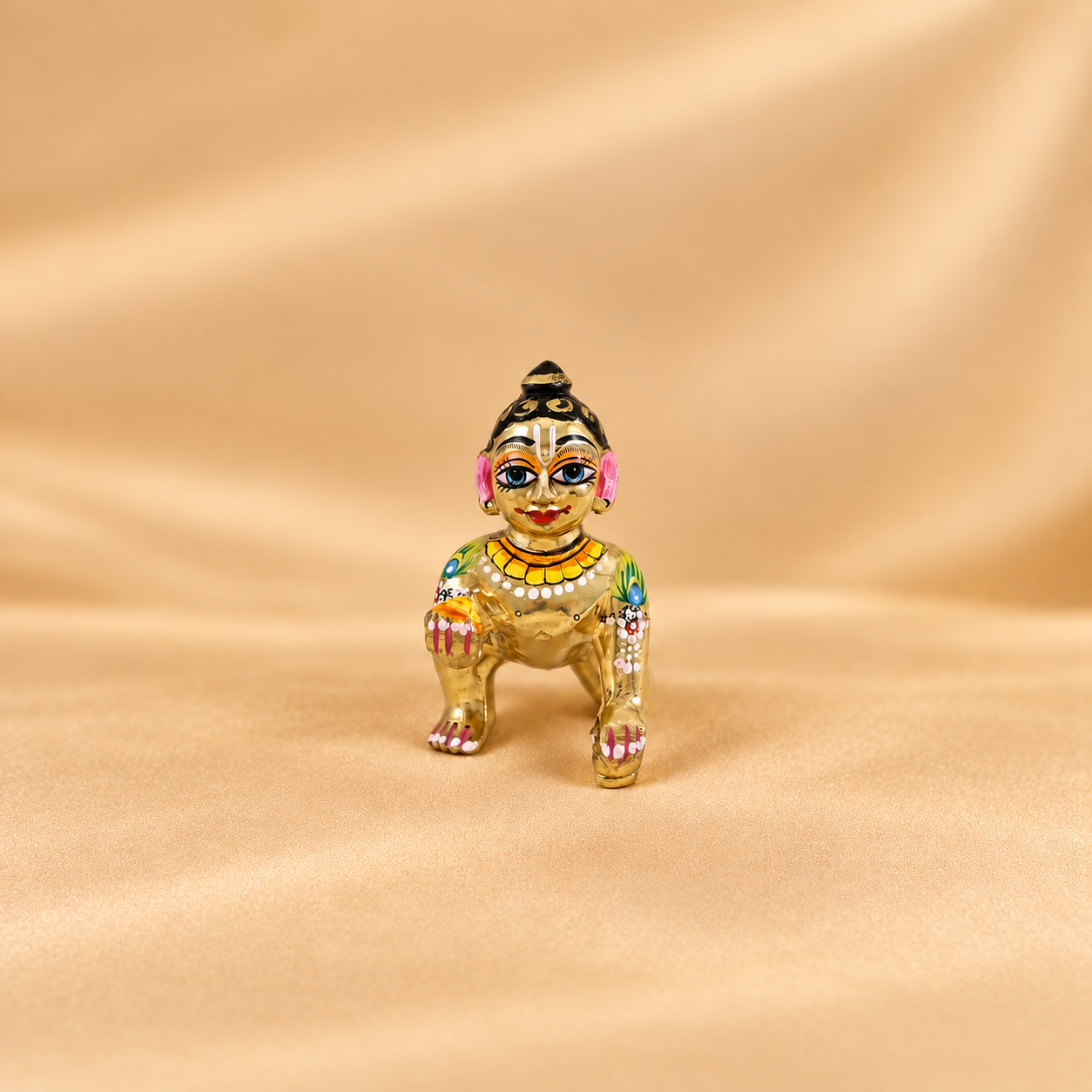 A small ornate figurine of a laddu gopal decorated with golden, pink, blue, green, and white details, standing on a beige satin fabric background.